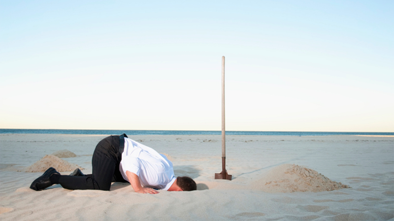 Man in wit overhemd steekt zijn hoofd in het zand op het strand, met een schop ernaast